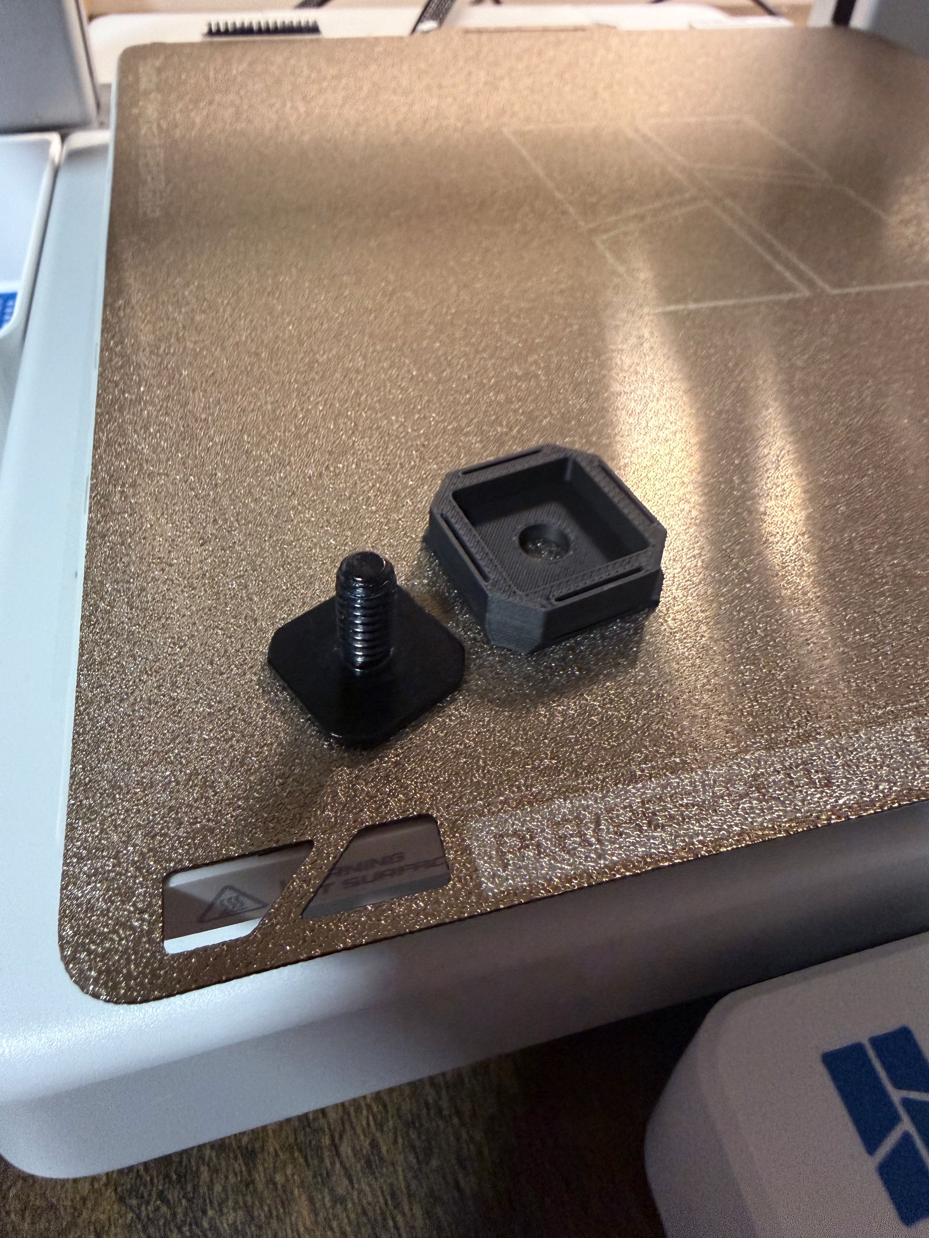 Photo of a 3D printer bed, with piece of metal with a tripod mount stuck to it, next to a 3D part that has a hole for that piece of metal.
