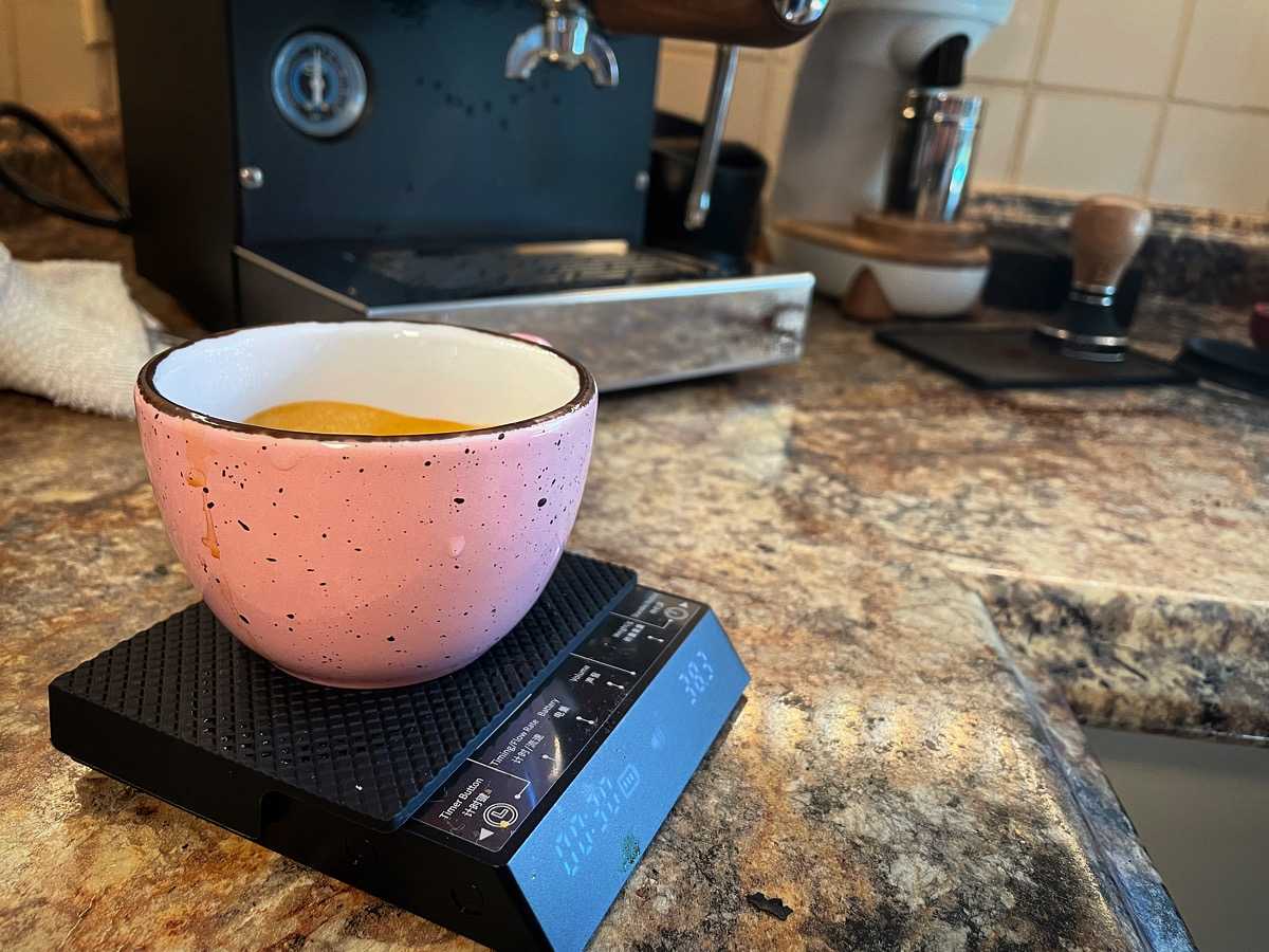 Photo of a small pink cup of espresso on a set of scales, with an espresso machine and other gear in the background